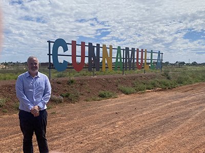 A bearded man in a blue business shirt and black pants stands in front of large colourful letters that spell Cunnamulla